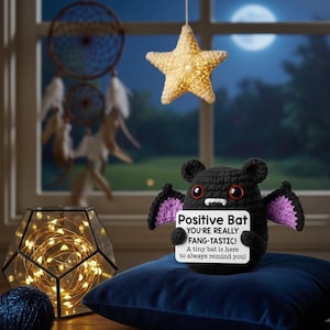 Op de afbeelding: Een zwarte gehaakte vleermuis met paarse vleugels en rode ogen houdt een bord vast met de tekst "Positive Bat, YOU'RE REALLY FANG-TASTIC! A tiny bat is here to always remind you!". De vleermuis zit op een blauw kussen.