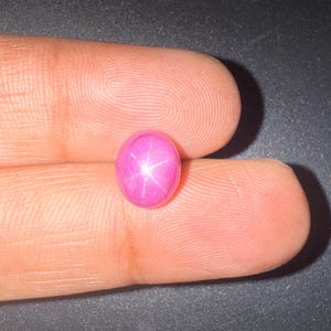 Pode incluir: Uma gema de safira estrela rosa de formato oval com um padrão de estrela de seis pontas distinto centrado na sua superfície cabochão lisa e polida. A gema exibe um tom magenta profundo e saturado com um efeito de asterismo claro.