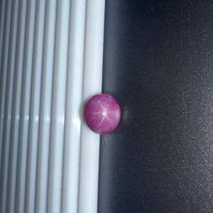 Könnte beinhalten: Ein runder, im Cabochon-Schliff gefertigter Sternrubin-Edelstein mit einem tiefen Magenta-Pink-Farbton. Der Stein zeigt einen ausgeprägten sechsstrahligen Sterneffekt, bekannt als Asterismus, der sich auf seiner polierten, gewölbten Oberfläche befindet.