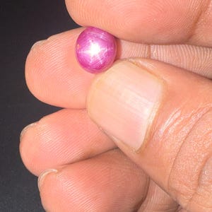 May include: A round, cabochon-cut star ruby gemstone with a vibrant magenta-pink hue. The stone displays a distinct six-rayed star effect, known as asterism, which appears as a bright white light reflection centred on the polished, smooth surface.