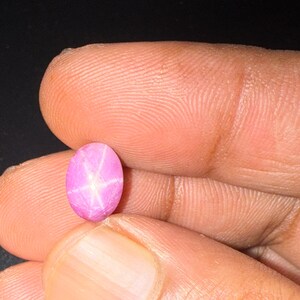 May include: An oval-shaped pink star sapphire gemstone featuring a distinct six-rayed star pattern visible on its smooth, polished surface. The gemstone displays a vibrant, translucent pink hue and is held in a vertical orientation.