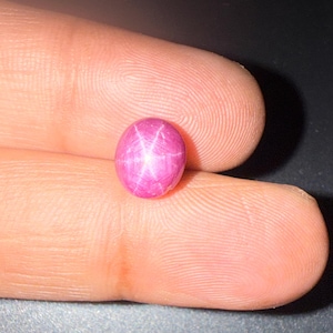Pode incluir: Uma pedra preciosa de rubi estrela de formato oval, polida, com um tom magenta-rosa vibrante e um padrão distinto de estrela de seis raios, conhecido como asterismo, visível na sua superfície lisa e abobadada de cabochão.