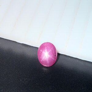 Puede incluir: Una piedra preciosa de forma ovalada, tallada en cabujón, en un tono rosa magenta vibrante. La piedra muestra un patrón de estrella de seis puntas distintivo en su superficie pulida y abovedada, característico del asterismo, con un aspecto liso.