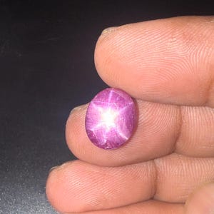 Può includere: Una gemma di rubino stellato a cabochon di forma ovale con una profonda tonalità magenta-rosa. La pietra presenta un distinto effetto a stella a sei raggi, noto come asterismo, che appare come un riflesso di luce bianca sulla superficie.