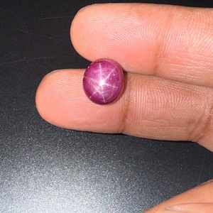 May include: A polished, oval-shaped cabochon gemstone with a deep magenta-red color. The surface of the stone displays a distinct six-rayed star pattern, known as asterism, which is centered on the dome of the smooth, translucent gemstone.