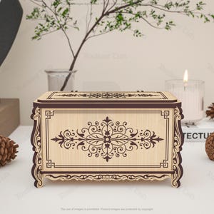 May include: A decorative wooden box with intricate floral and scrollwork designs. The box is light-colored with dark brown accents and a rectangular shape. The design is symmetrical and features a central floral motif. The box is displayed with pinecones and a candle.