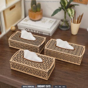 Puede incluir: Tres cajas rectangulares para pañuelos de madera de color marrón claro con intrincados patrones cortados con láser en la parte superior y los lados. Cada caja contiene un pañuelo blanco, con diseños que incluyen motivos de ondas geométricas.