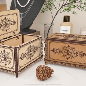 May include: Two wooden decorative boxes with ornate floral designs. One box is open, revealing the interior, while the other is closed. The boxes are light brown with dark brown trim and detailing. A pinecone sits in front of the boxes.