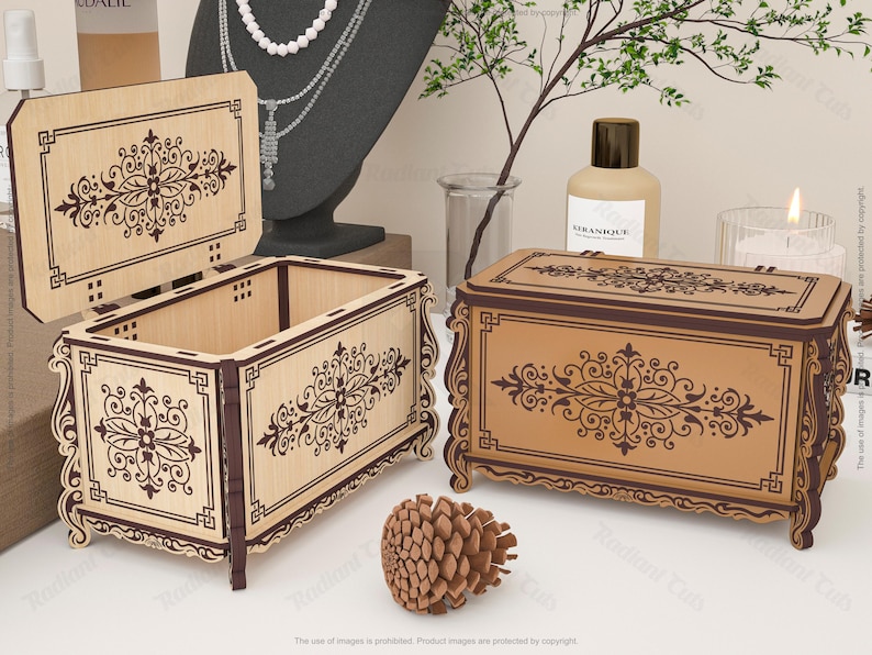 May include: Two wooden decorative boxes with ornate floral designs. One box is open, revealing the interior, while the other is closed. The boxes are light brown with dark brown trim and detailing. A pinecone sits in front of the boxes.