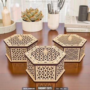 May include: Three laser-cut wooden hexagon boxes with intricate geometric patterns. The boxes have a light wood color with dark brown trim. The lids feature a decorative teardrop-shaped accent. The boxes are displayed on a brown surface.