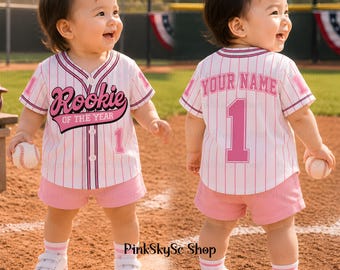 May include: A baby wearing a pink and white baseball jersey with the text "Rookie of the Year" and the number 1. The baby is also wearing pink shorts, white shoes, and holding a baseball. The jersey has pink stripes.