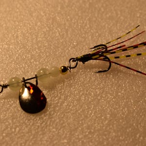 May include: A fishing lure with a black hook, a reflective teardrop-shaped blade, and a series of colorful feathers. The lure also features a small gold bead and several translucent beads.