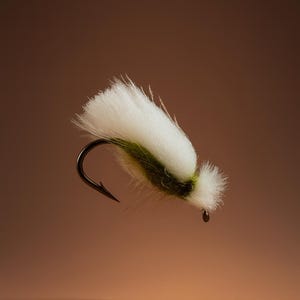 May include: A close-up of a fly fishing lure. The lure features a white, fluffy body with a green band and a black hook. The background is a gradient of brown.