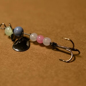 May include: A fishing lure with a silver blade and a series of colorful beads in light green, blue, white, and pink. The lure has a silver hook and is designed for catching fish. The lure is on a brown background.