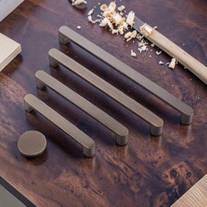 May include: A collection of bronze-toned cabinet hardware, including four rectangular pulls of varying lengths and a round knob. The hardware is arranged on a dark wooden surface, with wood shavings and a carving tool visible in the background.