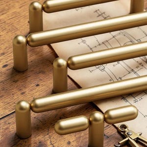 May include: Close-up of several gold-colored cabinet pulls arranged on a wooden surface. The pulls have a cylindrical bar design with rounded ends. A vintage blueprint and a brass compass are also visible.