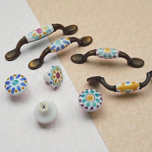 Può includere: Una collezione di ferramenta decorativa per mobili, tra cui maniglie e pomelli. Le maniglie hanno una base curva color bronzo con un centro in ceramica bianca con motivi floreali colorati. I pomelli sono rotondi con disegni simili.