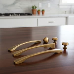 Peut inclure: Ensemble de quincaillerie de placard dorée, comprenant trois poignées incurvées et trois boutons. Le matériel est disposé sur une surface en bois foncé, mettant en valeur le design et la finition. Idéal pour la rénovation de placards et tiroirs.