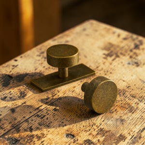 Può includere: Due pomelli e maniglie per mobili in ottone antico su una superficie di legno stagionato. Un pomello è rotondo con un piccolo stelo, l'altro ha una base rettangolare con una parte superiore circolare. L'hardware ha un aspetto vintage e invecchiato.