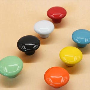May include: A collection of colorful ceramic cabinet knobs in various shades, including red, white, black, blue, yellow, green, and orange. Each knob has a rounded top and a short, cylindrical base. The knobs are arranged on a light brown surface.