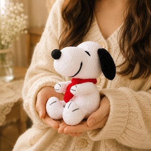 May include: A white plush Snoopy doll with black ears and a black nose, wearing a red scarf. The doll is held in someone's hands. The background includes a vase of white flowers and a lit candle.