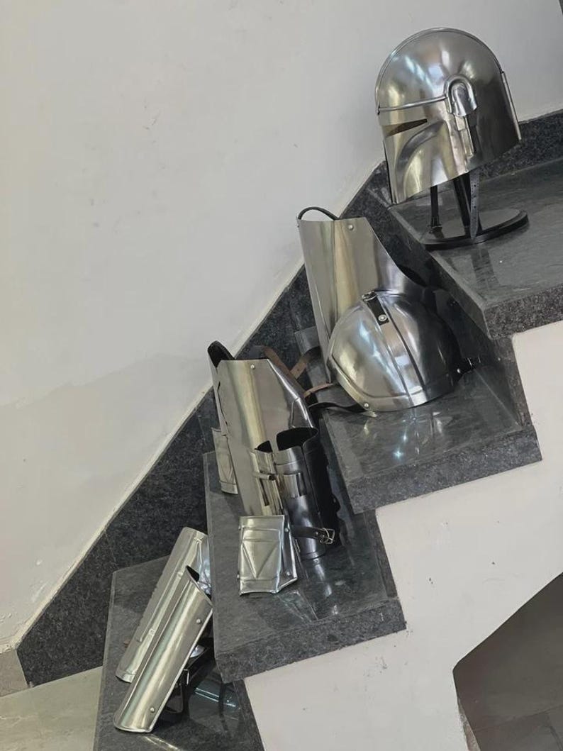 May include: A collection of shiny, silver-colored Mandalorian armor pieces, including a helmet, chest plate, and leg guards. The armor is displayed on a set of stairs with a dark gray surface.