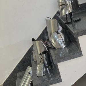 May include: A collection of shiny, silver-colored Mandalorian armor pieces, including a helmet, chest plate, and leg guards. The armor is displayed on a set of stairs with a dark gray surface.