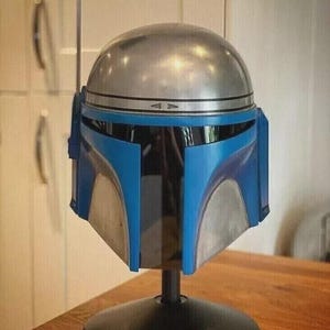 May include: A Mandalorian helmet replica, featuring a silver dome and blue accents. The helmet is mounted on a black stand, showcasing its iconic design. The helmet has a silver band around the middle and a blue visor.