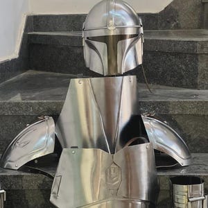 May include: A complete set of silver-colored Mandalorian armor, including a helmet, chest plate, shoulder pads, and other components. The armor is displayed on a staircase, showcasing its polished metal finish and intricate design.