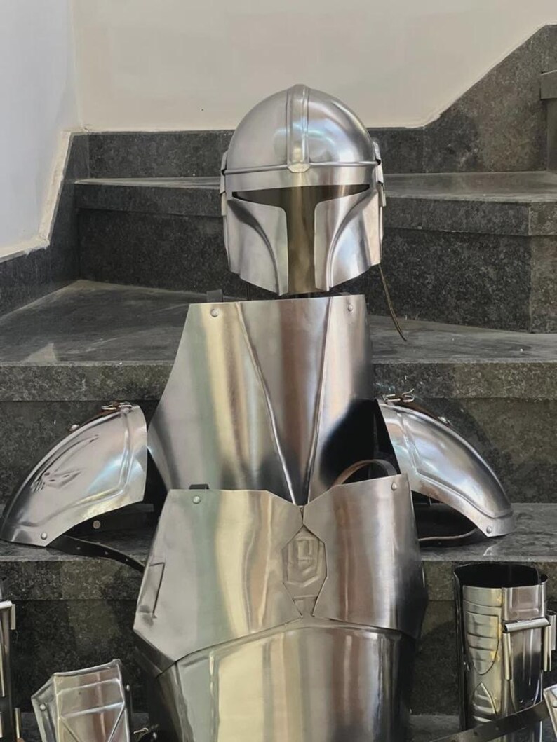 May include: A complete set of silver-colored Mandalorian armor, including a helmet, chest plate, shoulder pads, and other components. The armor is displayed on a staircase, showcasing its polished metal finish and intricate design.