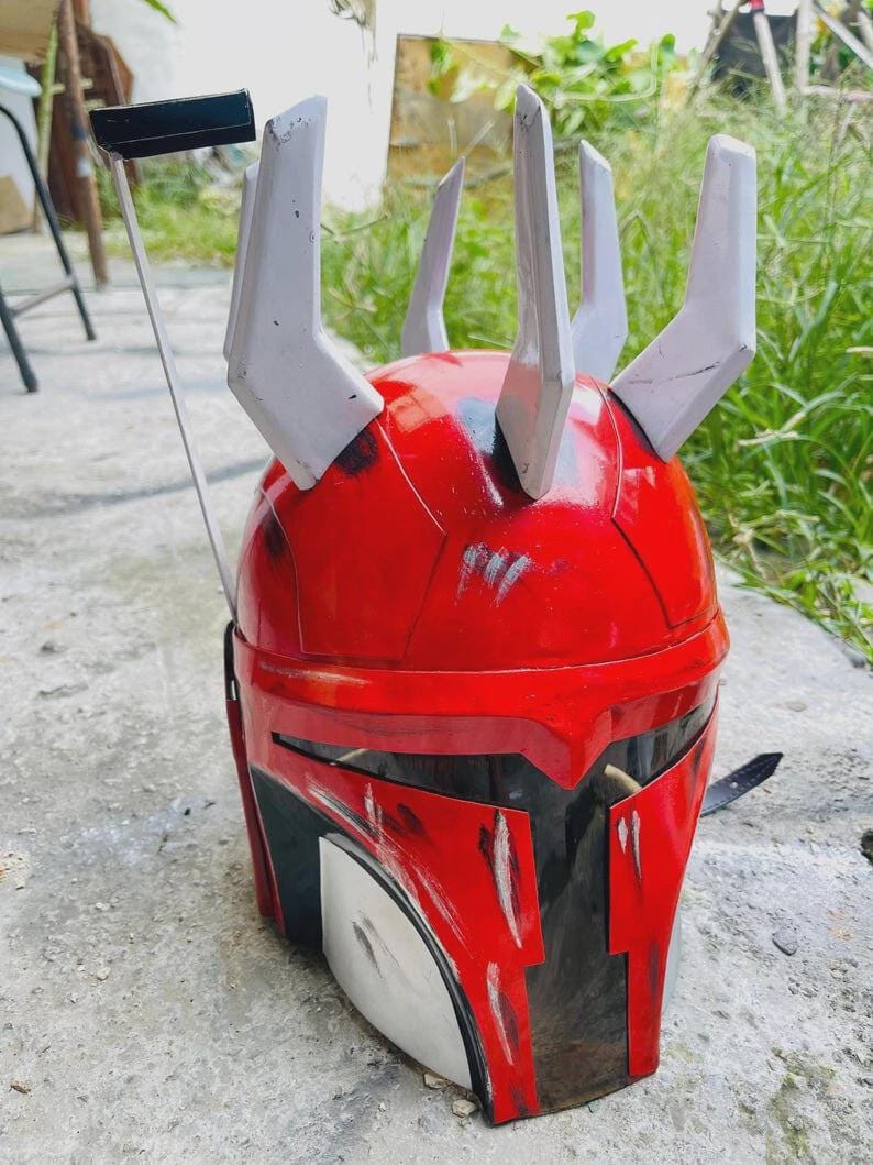 May include: A red and black helmet with white accents, featuring a unique design with multiple upward-pointing projections. The helmet appears to be made of a hard material and has a glossy finish. The design includes a visor area and a strap.