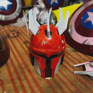 May include: A red and silver Mandalorian helmet with white details and a T-shaped visor. The helmet has a worn look, with Captain America shields visible in the background.