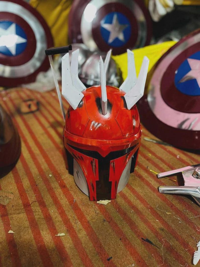 May include: A red and silver Mandalorian helmet with white details and a T-shaped visor. The helmet has a worn look, with Captain America shields visible in the background.
