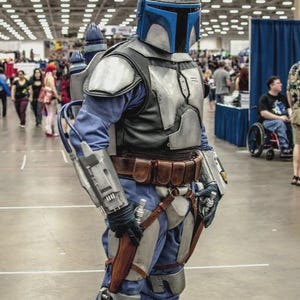 May include: A person in a detailed Mandalorian costume, featuring a silver and blue helmet, armor, and a jetpack. The costume includes a blue jumpsuit, brown belt, and various weapon accessories. The setting appears to be a convention hall.