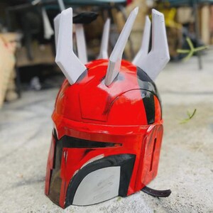 May include: A red and black helmet with white details, showcasing a unique design with several upward-pointing spikes. The helmet has a shiny surface and a dark visor area, possibly inspired by science fiction or fantasy.