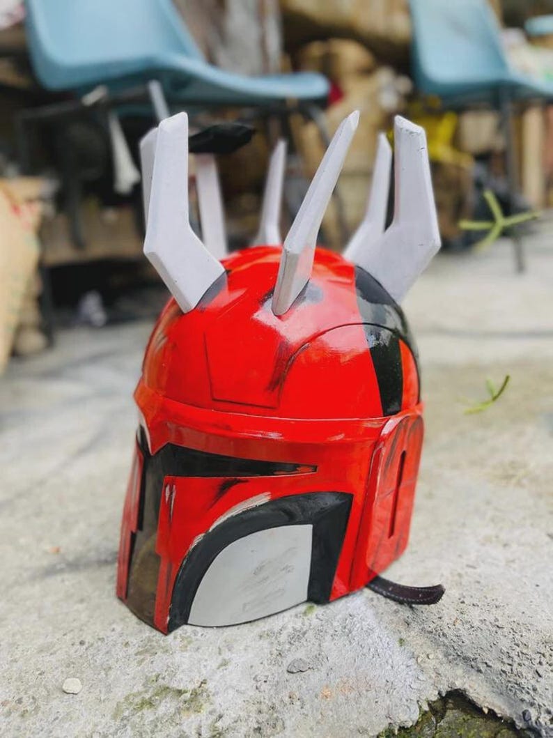 May include: A red and black helmet with white details, showcasing a unique design with several upward-pointing spikes. The helmet has a shiny surface and a dark visor area, possibly inspired by science fiction or fantasy.