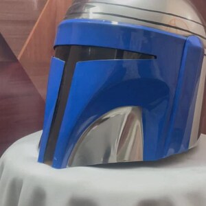 May include: A metallic silver and blue helmet with a visor. The helmet has a rounded top and a blue faceplate with black accents. The helmet is displayed on a white surface.