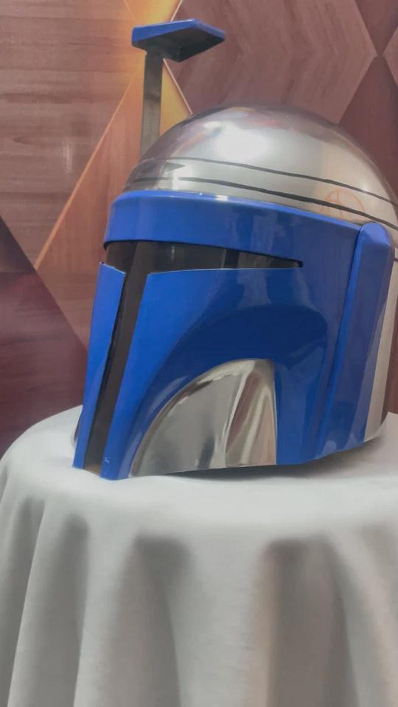 May include: A metallic silver and blue helmet with a visor. The helmet has a rounded top and a blue faceplate with black accents. The helmet is displayed on a white surface.