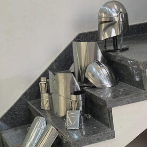 May include: A collection of shiny, silver-colored Mandalorian armor pieces, including a helmet, chest plate, and various arm and leg guards. The armor is displayed on a set of gray granite steps, showcasing its reflective surface and intricate design.