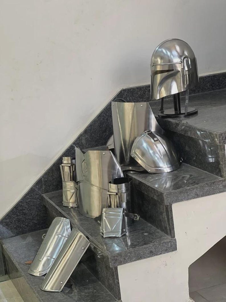 May include: A collection of shiny, silver-colored Mandalorian armor pieces, including a helmet, chest plate, and various arm and leg guards. The armor is displayed on a set of gray granite steps, showcasing its reflective surface and intricate design.