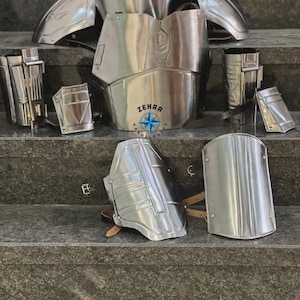 May include: A collection of polished silver metal armor pieces, including a chest plate, shoulder guards, and arm and leg protection. The armor is displayed on a set of gray steps. The text "ZEHRA HANDICRAFTS" is visible.