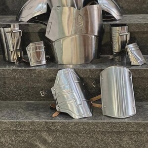May include: A set of polished silver metal armor pieces, including a chest plate, shoulder guards, arm and leg pieces, and other components. The armor is displayed on a set of gray steps, showcasing its reflective surface and intricate design.