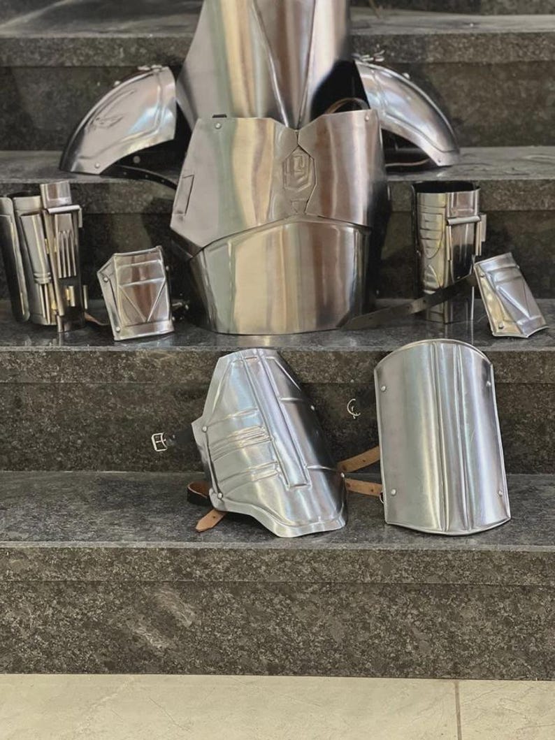 May include: A set of polished silver metal armor pieces, including a chest plate, shoulder guards, arm and leg pieces, and other components. The armor is displayed on a set of gray steps, showcasing its reflective surface and intricate design.