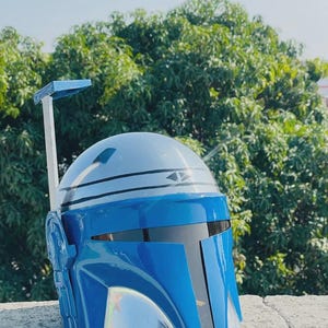 May include: A blue and silver Mandalorian helmet with a T-shaped visor. The helmet has a silver rangefinder and a blue and silver color scheme. The helmet is made of a glossy material and is likely a costume piece.