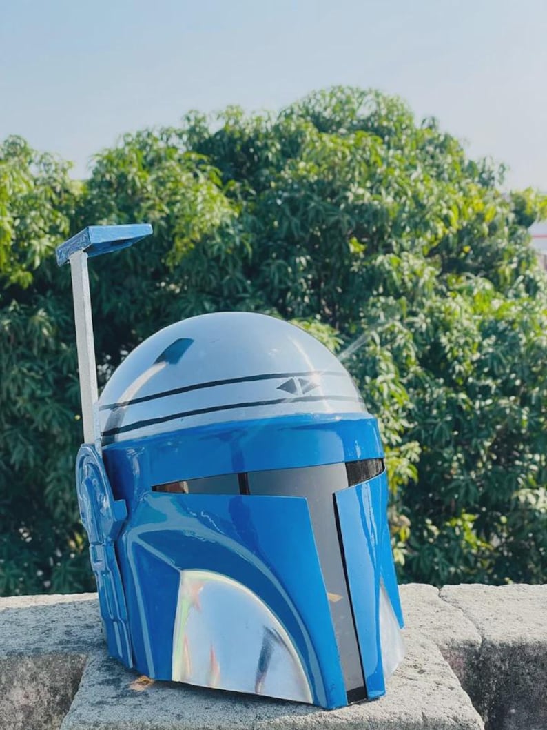 May include: A blue and silver Mandalorian helmet with a T-shaped visor. The helmet has a silver rangefinder and a blue and silver color scheme. The helmet is made of a glossy material and is likely a costume piece.