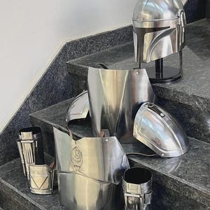 May include: A complete set of shiny, silver-colored Mandalorian armor, including a helmet, chest plate, shoulder pads, and leg guards. The armor is displayed on a set of stairs, showcasing its intricate design and metallic finish.
