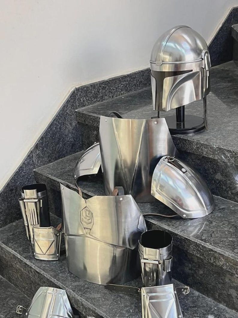 May include: A complete set of shiny, silver-colored Mandalorian armor, including a helmet, chest plate, shoulder pads, and leg guards. The armor is displayed on a set of stairs, showcasing its intricate design and metallic finish.