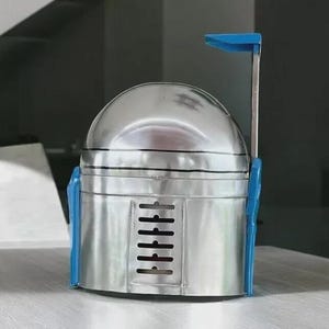 May include: A metallic silver helmet with blue accents, reminiscent of a Mandalorian helmet. The helmet features a rounded top, a rectangular lower section with vertical vents, and a blue antenna-like structure extending upwards. The design is a unique, handcrafted item.