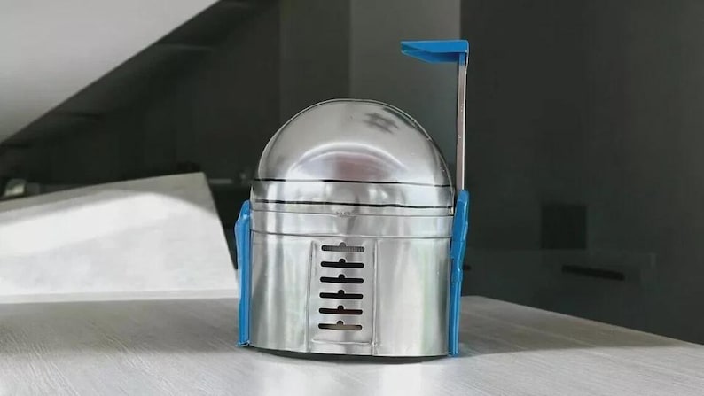 May include: A metallic silver helmet with blue accents, reminiscent of a Mandalorian helmet. The helmet features a rounded top, a rectangular lower section with vertical vents, and a blue antenna-like structure extending upwards. The design is a unique, handcrafted item.