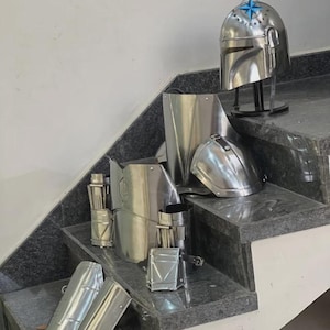 May include: A collection of shiny, silver-colored Mandalorian armor pieces, including a helmet, chest plate, and arm guards. The helmet is displayed on a black stand. The armor is arranged on a set of gray steps.